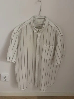 Randig linneskjorta från Pull&Bear - Säljer en vit skjorta från Pull&Bear med tunna vertikala ränder och bröstficka. Skjortan är tillverkad i linnemix, har korta ärmar och klassisk krage. Perfekt för en avslappnad och stilren look.