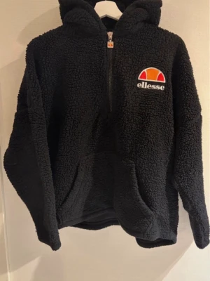Ellesse teddytröja - Svart huvtröja från Ellesse med dragkedja framtill och en känguruficka. Tröjan har en huva och långa ärmar. Ellesse-logotypen är placerad på bröstet. Rökfritt hem