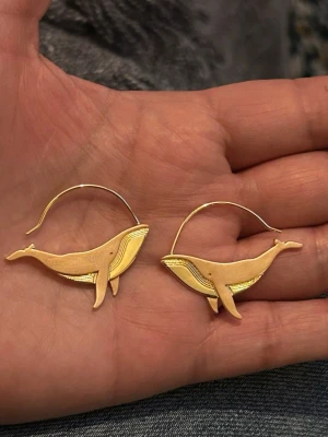 Guldiga örhängen med valmotiv - örhängen i form av valar, tillverkade i metall med en guldig finish. De har en stilren och lekfull design som verkligen sticker ut. Perfekta för dig som gillar havsinspirerade accessoarer och vill ha något annorlunda.