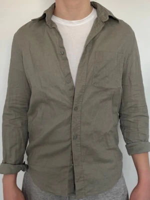 Olivgrön linneskjorta med bröstficka - Säljer en olivgrön skjorta i linne med klassisk krage och bröstficka. Skjortan har långa ärmar som kan vikas upp och stängs med knappar framtill. Perfekt för en avslappnad och stilren look.