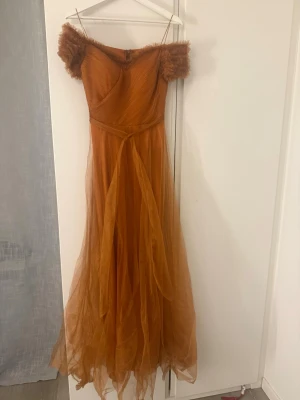 Lång orange offshoulder klänning - Snygg långklänning i orange med offshoulder ärmar och tunna axelband. Klänningen har ett lager av tunn tyll som ger ett drömmigt intryck och volym. Perfekt för dig som vill sticka ut med en unik och elegant look.