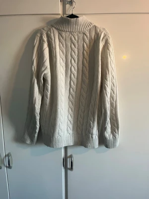 Vit kabelstickad tröja Polo Ralph Lauren - Kabelstickad vit tröja från Polo Ralph Lauren med halv dragkedja och hög krage. Tröjan har ribbade muddar och nederkant samt den klassiska broderade loggan på bröstet. Perfekt för dig som gillar en clean och tidlös stil. Jag är 184 cm lång vikt 68 kg och den sitter perfekt på mig.