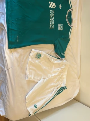 Liverpool FC bortaställ Adidas - Säljer ett Liverpool FC bortaställ från Adidas med grön matchtröja och vita shorts. Tröjan har klubbmärke, sponsorlogga och klassiska Adidas-ränder på axlarna. Shortsen är vita med gröna detaljer och klubbmärke. Perfekt för fotbollsträning eller match!