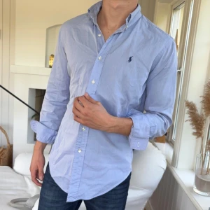 Ljusblå skjorta från Ralph Lauren - Säljer en klassisk ljusblå skjorta från Ralph Lauren i bomull. Skjortan har lång ärm, knappar framtill och den ikoniska broderade loggan på bröstet. Passformen är normal (classic fit) och materialet är mjukt och skönt. Perfekt till jeans eller chinos.