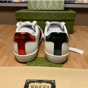 Gucci Ace sneakers med orm-motiv - Vita Gucci Ace sneakers i läder med ikoniska broderade ormar på sidorna. Baksidan har metallicröd detalj på ena skon och metallicgrön på den andra. Klassisk låg modell med vita snören och gummisula. Perfekt för dig som vill sticka ut med lyxig streetstyle.