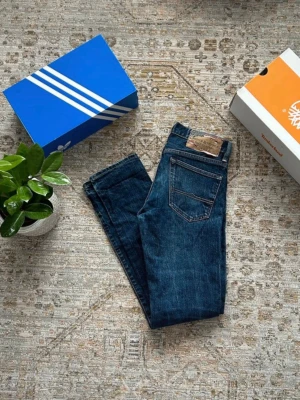 Ralph Lauren Jeans - Säljer ett par klassiska blå jeans från Ralph Laren Denim & Supply med smal passform och fem fickor. Jeansen har en mörk tvätt och detaljer i kopparfärg på knappar och nitar. I otroligt fint skick. Perfekta för dig som gillar tidlös stil och vill ha ett par jeans som funkar till allt. Storlek W29 L32. Pris kan alltid diskuteras✅.