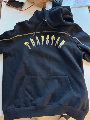 TRAPSTAR hoodie ”Central Cee edition” - Ordentlig och bra kvalité. Tjockt äkta material. Säljer en svart hoodie från Trapstar med stor gul och vit logga på bröstet. Hoodien har huva med dragsko, magficka och kontrastsömmar längs ärmarna. Perfekt för dig som gillar streetwear och vill sticka ut med en snygg detalj på outfiten.
