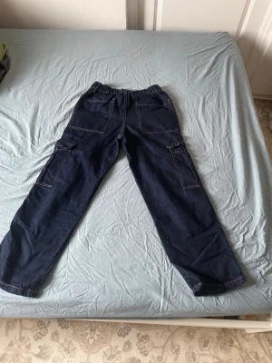 Mörkblå cargobyxor med fickor - Säljer ett par mörkblå cargobyxor i denim med breda ben och stora sidofickor. Byxorna har resår i midjan och kontrastsömmar för en cool look. Perfekta för dig som gillar streetwear och praktiska detaljer.