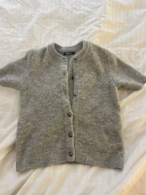 Grå stickad kortärmad topp Ginatricot - Mysig grå stickad topp från Ginatricot med korta ärmar och rund hals. Toppen har knappar framtill och ribbade kanter vid ärmslut och nederkant. Perfekt att styla lager på lager eller bära som den är.