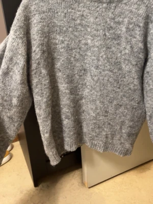 Grå stickad tröja - Mysig grå stickad tröja med rund hals och avslappnad passform. Tröjan har långa ärmar och ribbade muddar vid ärmslut och nederkant. Perfekt för kyliga dagar och enkel att matcha med jeans eller kjol.