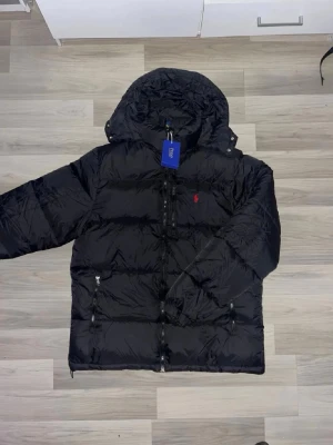 Svart pufferjacka från Polo Ralph Lauren - Snygg svart pufferjacka från Polo Ralph Lauren med huva och dragkedja framtill. Jackan har två framfickor med dragkedja och den klassiska röda loggan broderad på bröstet. Perfekt för kalla vinterdagar och riktigt skön att bära.