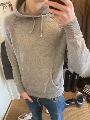 Grå hoodie med ficka framtill - Säljer en stilren grå hoodie med huva och dragsnören. Hoodien har en stor magficka och ribbade muddar vid ärmslut och nederkant. Perfekt för en avslappnad look och passar till jeans eller joggers.
