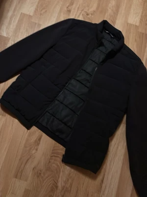 Cardigan, ZARA - Säljer en svart pufferjacka med quiltade paneler och hel dragkedja framtill. Jackan har en klassisk, rak passform och är tillverkad i ett syntetmaterial som håller värmen. Perfekt för kalla vinterdagar och har en stilren look.