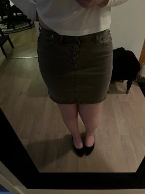 Kort militärgrön kjol med knappar - Säljer en kort militärgrön kjol med knappar framtill och klassiska fickor. Kjolen har en enkel och stilren design som passar perfekt till en avslappnad look. Materialet känns som bomull eller denim och den har en normal passning.