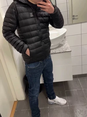 Svart pufferjacka från Haglöfs - Säljer en svart pufferjacka från Haglöfs med huva och dragkedja framtill. Jackan har en quiltad design och två sidofickor. Perfekt för kalla dagar och riktigt skön att bära. Klassisk och stilren look som funkar till det mesta.