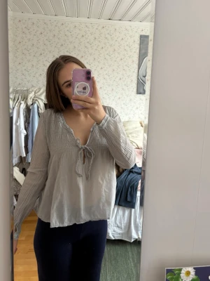 Ljusgrå blus med knytning fram - Jättefin ljusgrå luftig blus med långärmad design och v-ringning med knytband framtill. Blusen har rynkade detaljer vid axlar och bröst. 