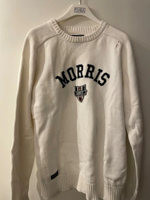 Vit stickad tröja från Morris - Vit stickad tröja från Morris med svart broderad logga och emblem på bröstet. Tröjan har rund halsringning, långa ärmar och ribbade muddar. Perfekt för en avslappnad och sportig look.