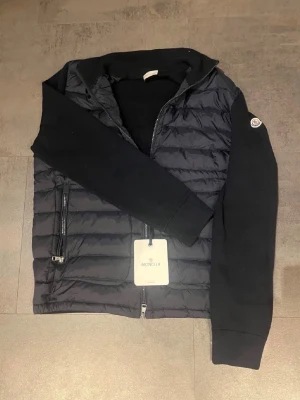 Svart dunväst från Moncler - Snygg svart dunväst från Moncler med quiltad front och två dragkedjefickor. Västen har en hög krage och Moncler-logga på ärmen. Materialet är en mix av dun och stickad bomull vilket ger en sportig och modern look. Storleken är XL men sitter som en L