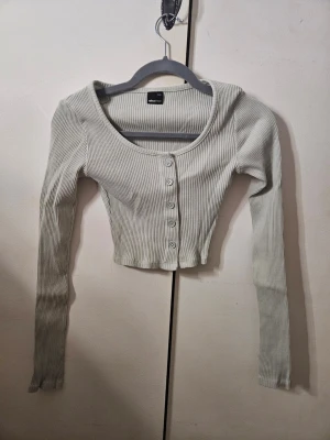 Ljusribbad croppad topp Gina Tricot - Superfin ljusgrön croppad topp från Gina Tricot med ribbad struktur och rundad halsringning. Toppen har långa ärmar och fem knappar framtill som ger en chill vibe. Endast använda ett fåtal gånger och i mycket bra skick. Nypris runt 150 kr. OBS. Tröjan ser mindre grön ut på bilderna än vad den är, se sista bild för en mer realistisk färg.