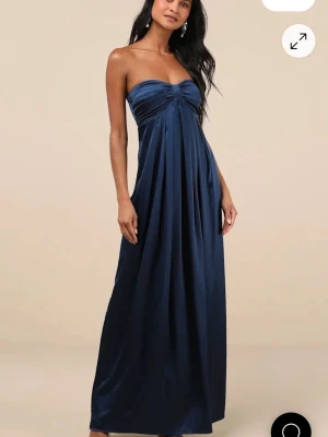 Balklänning - Elegant marinblå långklänning med axelbandslös design och draperad överdel. Klänningen har ett flödande fall och är tillverkad i ett glansigt material som ger en lyxig känsla. Perfekt för speciella tillfällen. Säljer pågrund av att den var för stor.
