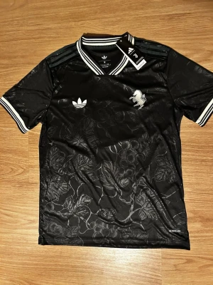 Svart Adidas fotbollströja Yildiz 10 - Snygg svart fotbollströja från Adidas med vita detaljer, blommönster och tryck på bröstet. På ryggen står det YILDIZ och nummer 10 i vitt. Tröjan har korta ärmar, ribbade muddar och klassiska Adidas-ränder på axlarna.