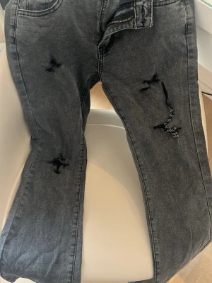 Svarta bootcut jeans med hål - Säljer ett par svarta jeans med slitningar och hål på benen. Jeansen har en klassisk femficksmodell och dragkedja i gylfen. Perfekta för en avslappnad och trendig look. Materialet är bomull med lite stretch för extra komfort.