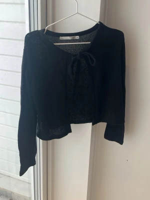 Svart cropped kofta med knytband - Trendig svart cropped kofta med långa ärmar och knytband framtill. Perfekt att slänga över ett linne eller klänning för en snygg lager-på-lager-look. Tunn och lättstickad för en avslappnad vibe.