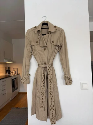Beige trenchcoat med nitar - En lite annorlunda och unik trenchcoat med bitar & snygga detaljer. Jackan är i mycket bra skick! Den är i storlek L men skulle säga att den är mer som en M. 