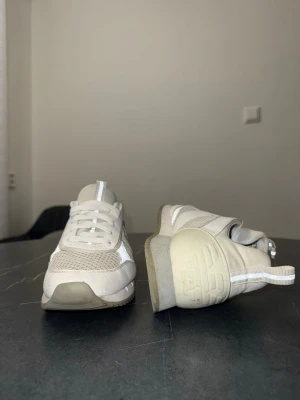Vita sneakers från EA7 Emporio Armani - Snygga vita sneakers från EA7 Emporio Armani med meshpaneler och detaljer i syntetläder. Skorna har vita snören, logga på sidan och dragflikar både fram och bak. Perfekta för en clean och sportig look.