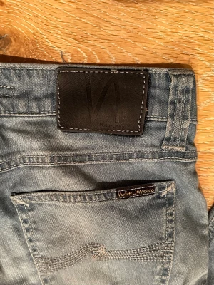 Blå slim fit jeans från Nudie Jeans - Säljer ett par blå slim fit jeans från Nudie Jeans med klassisk femficksmodell och svarta läderpatch bak. Jeansen har en snygg tvättad look och subtila slitningar. Perfekta för dig som gillar en avslappnad men stilren vibe.