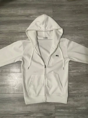 Vit zip hoodie från Lacoste - Snygg vit hoodie från Lacoste med dragkedja framtill och klassisk krokodil-logga på bröstet. Hoodien har två stora fickor, justerbar huva med snören och ribbade muddar. Tillverkad i mjukt bomullsmaterial som känns skönt mot huden.