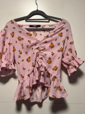 Rosa blommig blus från Bik Bok - Söt rosa blus från Bik Bok med blommigt mönster i orange och grönt. Blusen har v-ringning, snörning framtill, volangdetaljer och korta ärmar med rynkade kanter. Perfekt för dig som gillar romantiska och trendiga toppar.