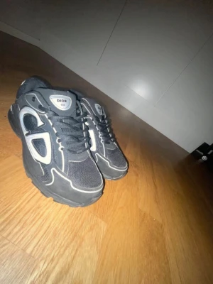 Svarta Dior sneakers med vita detaljer - Snygga svarta sneakers från Dior med vita detaljer och logga på innersulan. Skorna har en klassisk låg profil och är tillverkade i textil och syntetmaterial. Perfekta för dig som vill ha en exklusiv och trendig look.