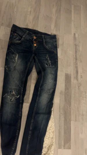 Mörkblå slitna jeans med knappar - Säljer ett par mörkblå jeans med slitningar och hål på benen. Jeansen har tre synliga kopparfärgade knappar och klassiska fem fickor. Snygg tvätt och coolt sliten look, perfekt för dig som gillar streetstyle.