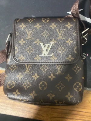 Louis Vuitton brun axelväska - Snygg brun axelväska från Louis Vuitton med klassiskt monogrammönster i beige och guldiga detaljer. Väskan har justerbar axelrem i brunt tyg och ett stort LV-emblem i metall på framsidan. Perfekt för dig som vill ha en ikonisk och trendig accessoar.