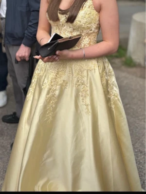 Ljusgul långklänning balklänning  - Elegant ljusgul balklänning med vackra spetsdetaljer upptill och vid midjan. Snörning bak för bästa passform. Klänningen har en utsvängd kjol och är ärmlös med en v-ringad urringning. Perfekt för bal eller student. Materialet har en glansig satinlook.