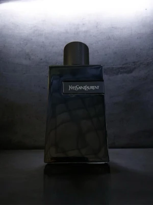 Yves Saint Laurent svart parfymflaska - Snygg parfymflaska från Yves Saint Laurent i svart glas med en stilren, fyrkantig form och metallisk rund kork med YSL-logga. Flaskan har en modern och exklusiv design som verkligen sticker ut på hyllan.