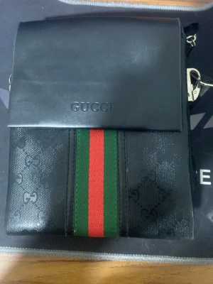 Svart axelväska från Gucci - Snygg svart axelväska från Gucci med klassiska gröna och röda ränder framtill. Väskan har lock med präglad logga och GG-mönster i materialet. Justerbar axelrem och detaljer i metall. Perfekt för dig som vill ha en stilren och ikonisk accessoar.
