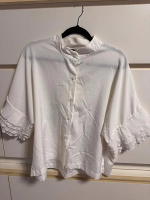 Vit blus med volangärm och knappar - Stilren vit blus med knäppning framtill och ståkrage. Blusen har vida trekvartsärmar med volangdetaljer och diskret struktur i tyget. Perfekt för en trendig och avslappnad look.