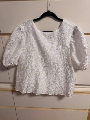 Vit puffärm blus med knyt i ryggen - Supersöt vit blus med puffärmar och struktur i tyget. Blusen har rund halsringning och en snygg knytning i ryggen som ger en extra detalj. Perfekt till jeans eller kjol för en trendig look.