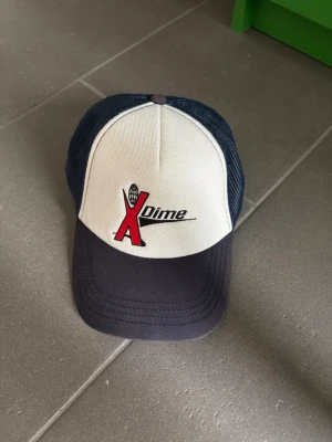 Dime truckerkeps blå och vit - Snygg truckerkeps från Dime med vit frontpanel och mörkblått mesh-tyg på sidor och bak. Kepsen har ett broderat X Dime-logo i rött och svart framtill samt böjd skärm i mörkblått. Justerbar baktill för perfekt passform.