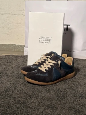 Maison Margiela Replica sneakers blå - Snygga Maison Margiela Replica sneakers i mörkblå mocka och läder med beige detaljer och gummisula. Klassisk low-top siluett och vita snören. Tillverkade i lamm- och kalvskinn för en lyxig känsla. Perfekta för dig som gillar stilrena och tidlösa sneakers.