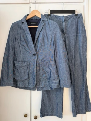 Blå denimkostym från Lene Sand jeans kavaj - Snygg blå denimkostym från Lene Sand med kavaj och matchande byxor. Kavajen har två stora fickor framtill, klassisk krage och knappstängning. Byxorna har raka ben och normal passform. Perfekt för dig som gillar en avslappnad men ändå stilren look.