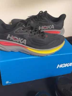 Svarta Hoka sneakers med färgglad sula - Snygga svarta sneakers från Hoka med mesh-överdel och en chunky sula i grått, gult och rött. Skorna har Hoka-logga på sidan och klassisk snörning. Perfekta för dig som gillar sportig stil och vill ha något bekvämt till vardags.