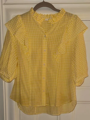 Gul rutig blus med volangdetaljer - Söt gul blus med rutmönster, volang vid axlar. Mycket bra skick. 