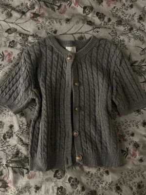 Grå kabelstickad kofta med kort ärm - Snygg grå kabelstickad kofta med korta ärmar och rund hals. Koftan har dekorativa guldfärgade knappar framtill och ribbade kanter. Perfekt att slänga över en t-shirt för en chill look.