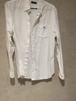 Vit skjorta från Gant med ficka - Klassisk vit skjorta från Gant med bröstficka och diskret logga. Skjortan har lång ärm, knappar framtill och en tidlös krage. Perfekt till både jeans och chinos för en clean look.