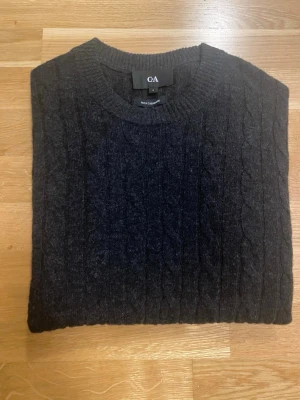 Mörkgrå kabelstickad tröja C&A - Snygg mörkgrå kabelstickad tröja från C&A med rund halsringning och långa ärmar. Tröjan har ribbade muddar vid ärmslut och nederkant, och är tillverkad i ett mjukt material som passar perfekt för kyligare dagar.