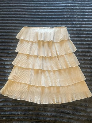 Plisserad klänning med volanger, beige - Säljer en superfin axelbandslös plisserad klänning i beige med flera lager volanger. Perfekt för dig som gillar en trendig och feminin look. Köpt sacond hand står ingen strl men passar en M