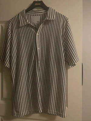 Randig skjorta från 157 i svart och vitt - Kortärmad skjorta från 157 med svartvita ränder och klassisk krage. Skjortan har knappar framtill och är gjord i ett luftigt material som passar perfekt till varmare dagar. Snygg och enkel att styla till olika outfits.